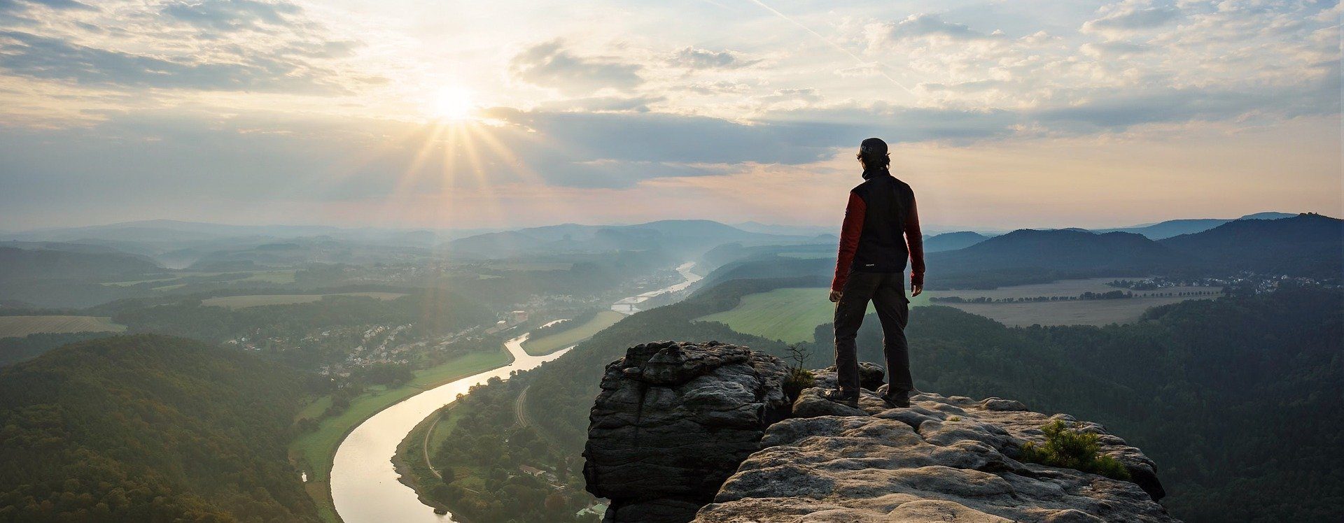The silhouette of a mountaineer in the sunset, standing on the edge of a cliff and looking down into the valley with a river.